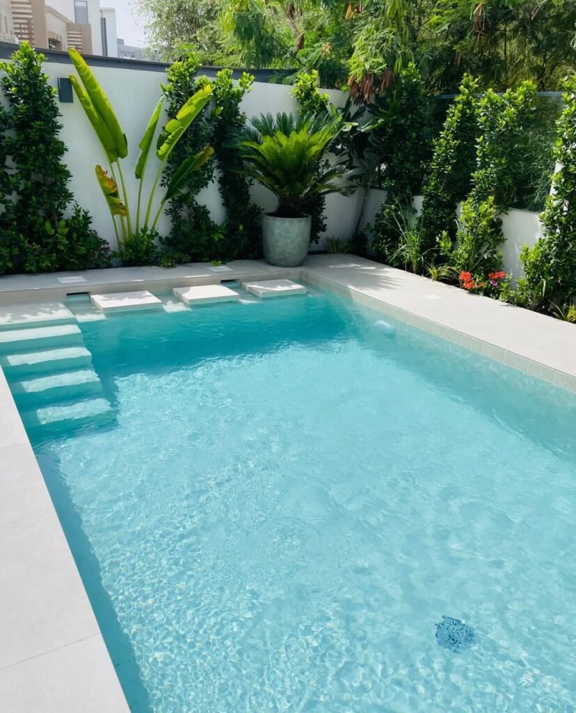A swimming pool with submerged entry steps, pale paving and planted borders along the boundary wall.