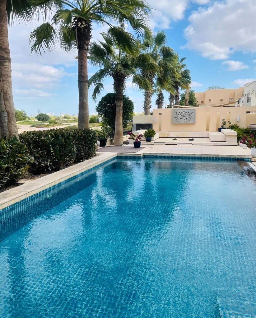 A rectangular swimming pool with paved edges, palm trees and seating at the far end.