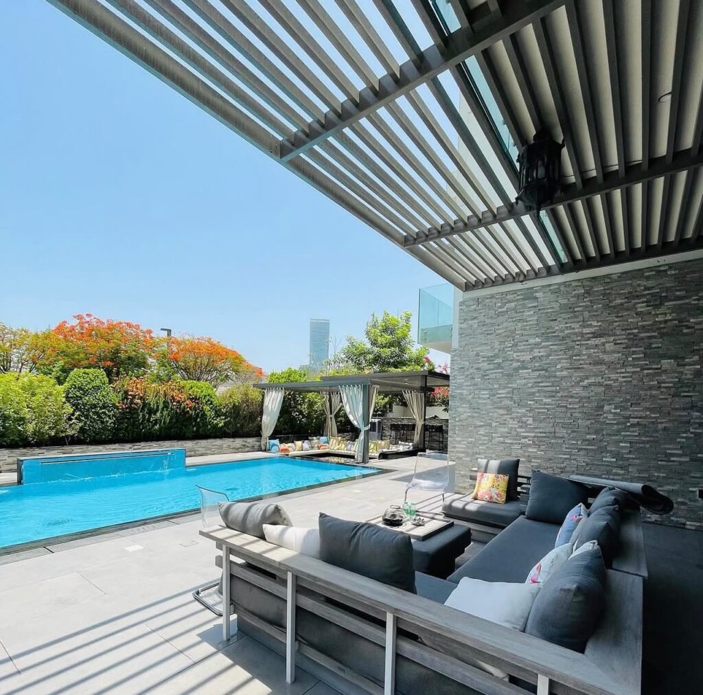 A poolside lounge beneath a slatted pergola, with a stone-clad feature wall, outdoor sofas and paved flooring.